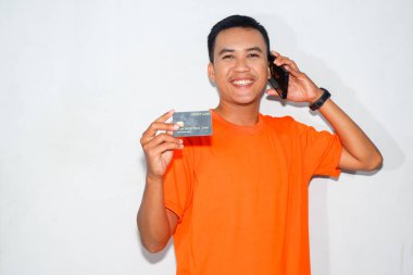 Asyalı adam turuncu tişört giyiyor ve gülümsüyor. Kredi kartı gösteriyor ve akıllı telefon kullanarak telefon ediyor.