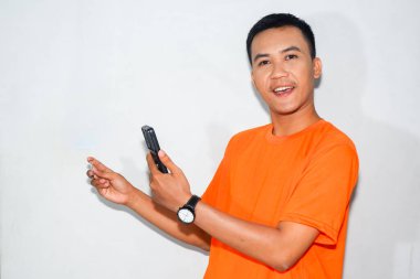 Asyalı adam turuncu tişört giyiyor, gülümsüyor, kredi kartı gösteriyor ve elinde akıllı telefonunu tutuyor.