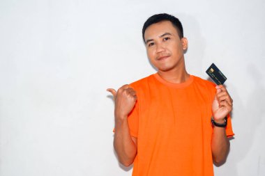 Asyalı adam turuncu gömlekli, mutlu bir ifadeyle kredi kartı gösteriyor ve baş parmağıyla işaret ediyor.