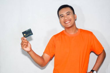 Asyalı adam turuncu tişört giyiyor ve gülümsüyor, siyah kredi kartı gösteriyor.