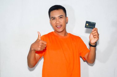 Turuncu tişört giyen Asyalı bir adam dostça bir ifadeyle kredi kartını gösteriyor ve başparmağını kaldırıyor.