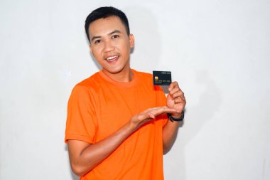 İki eliyle kredi kartı gösteren turuncu tişörtlü Asyalı bir adam.