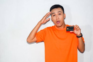 Turuncu tişörtlü Asyalı adam şaşırmış bir ifadeyle kredi kartı gösteriyor ve kafa hareketi yapıyor.