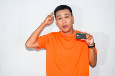 Turuncu tişört giyen kafası karışmış Asyalı bir adam. Kafasını kaşırken kredi kartını gösteriyor.