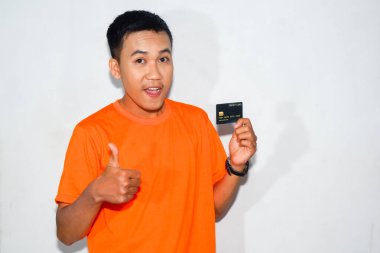 Dost canlısı, turuncu tişörtlü, kredi kartı gösteren ve baş parmak işareti yapan Asyalı bir adam..