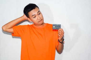 Kafası karışmış turuncu tişört giyen Asyalı bir adam kredi kartına bakarken kafasını tutuyor.