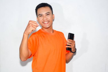 Dost canlısı, turuncu tişörtlü, elinde akıllı bir telefonla kutlama jesti yapan Asyalı bir adam. Başarı kavramı, başarı, çevrimiçi zafer veya olumlu haber