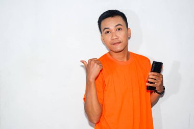 Dost canlısı, turuncu tişörtlü, akıllı bir telefon tutan ve baş parmağıyla yanlamasına işaret eden Asyalı bir adam. Gösterme, bilgi paylaşma, teknoloji kullanımı veya iletişim kavramı