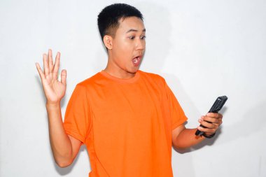 Turuncu tişört giyen Asyalı bir adam akıllı telefonuna bakıyor. Şok kavramı, beklenmedik haberler, dijital iletişim veya çevrimiçi tepki