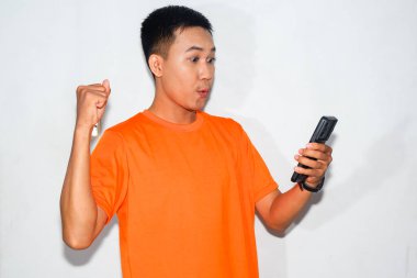 Turuncu tişört giyen mutlu Asyalı adam akıllı telefonuna sıkılmış yumruğuyla bakıyor. Heyecan, başarı, başarı veya pozitif dijital deneyim kavramı
