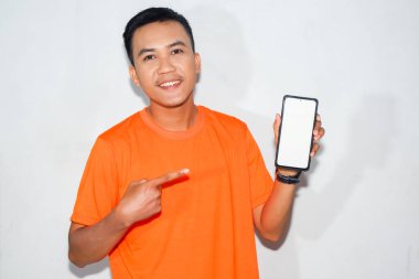 Turuncu tişörtlü arkadaş canlısı Asyalı adam akıllı telefonunun boş ekranını işaret ediyor..