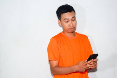 Hayal kırıklığına uğramış Asyalı bir adam turuncu tişört giyiyor, elinde akıllı bir telefonla ona bakıyor. Hayal kırıklığı, teknik sorun veya olumsuz dijital deneyim kavramı