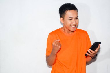 Turuncu tişört giyen mutlu Asyalı adam akıllı telefonuna bakıyor ve zafer jesti yapıyor. Başarı kavramı, başarı, kutlama veya pozitif dijital deneyim