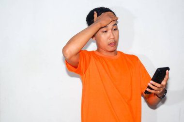 Hüsrana uğramış Asyalı adam turuncu tişört giyiyor, akıllı telefonuna bakıyor ve facepaling yapıyor. Hayal kırıklığı, dijital sorunlar, teknik sorunlar veya çevrimiçi hayal kırıklığı kavramı