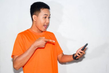 Turuncu tişört giyen enerjik Asyalı adam parmağıyla akıllı telefon ekranını işaret ediyor. Dijital etkileşim ve teknoloji kullanımı kavramı
