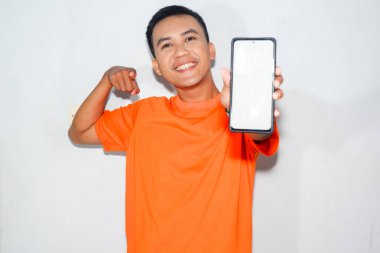 Asyalı adam gülümsüyor, turuncu tişört giyiyor, akıllı telefonun beyaz ekranını işaret ediyor. Teknoloji kavramı, mobil etkileşim veya akıllı telefon kullanımı