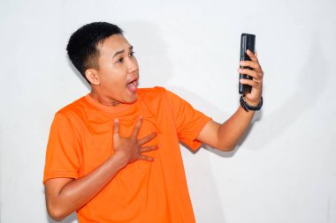 Turuncu tişört giyen Asyalı bir adam akıllı telefonun beyaz ekranına bakıyor ve göğsünü tutuyor. Şok, sürpriz veya beklenmedik dijital içerik kavramı