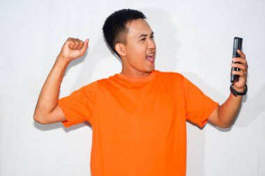 Turuncu tişört giyen Asyalı bir adam akıllı telefonun beyaz ekranına bakıyor ve zafer jesti yapıyor. Başarı, kutlama veya başarı kavramı