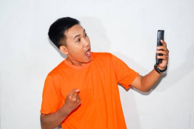 Turuncu tişört giyen Asyalı bir adam akıllı telefonun beyaz ekranına bakıyor ve zafer jesti yapıyor. Başarı, kutlama veya başarı kavramı