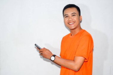 Şaşırmış bir suratı olan Asyalı bir adam, turuncu bir tişört giyiyor, akıllı bir telefon tutuyor ve kameraya bakıyor, yandan bakıyor. Sürpriz, dijital etkileşim ya da beklenmeyen tepki kavramı