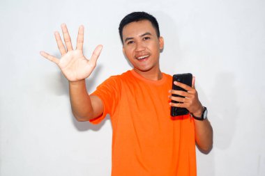 Turuncu tişört giyen, elinde akıllı telefon tutan ve el sallayan dost canlısı Asyalı bir adam. Selamlama, iletişim veya dijital etkileşim kavramı