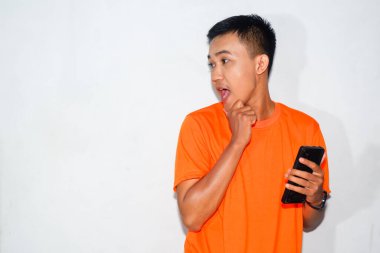 Dost canlısı, turuncu tişörtlü, elinde akıllı bir telefon olan Asyalı bir adam yan tarafa bakıyor. Merak, tefekkür ya da dijital etkileşim kavramı