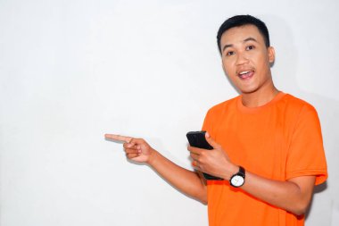 Dost canlısı, turuncu tişörtlü, akıllı telefon tutan ve parmağıyla kenarı gösteren Asyalı bir adam. Yön kavramı, dijital etkileşim veya rehberlik