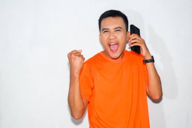 Turuncu tişört giyen neşeli Asyalı adam telefonda konuşuyor ve yumruk hareketi yapıyor. Heyecan, kutlama ya da pozitif iletişim kavramı