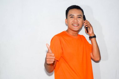 Dost canlısı, turuncu tişörtlü, telefonda konuşan ve baş parmak işareti yapan Asyalı bir adam. Onay, iletişim veya olumlu etkileşim kavramı
