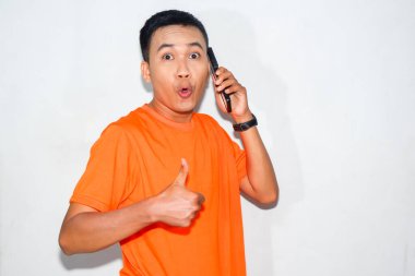 Turuncu tişört giyen neşeli Asyalı adam telefonda konuşuyor ve baş parmağını kaldırıyor. Onay, mutluluk veya olumlu iletişim kavramı