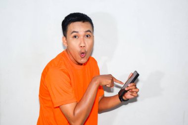 Turuncu tişört giyen komik Asyalı adam akıllı telefonunu işaret ediyor. Mizah anlayışı, teknoloji, ya da şaşırtma ve eğlendirme