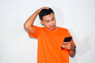 Turuncu tişört giyen kafası karışmış Asyalı adam akıllı telefonuna bakıyor. Teknolojiyi kullanırken kafa karışıklığı, hayal kırıklığı veya belirsizlik kavramı
