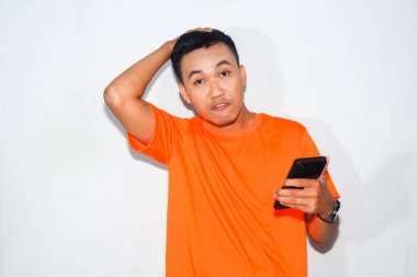 Turuncu tişört giyen kafası karışmış Asyalı adam akıllı telefonuna bakıyor. Teknolojiyi kullanırken kafa karışıklığı, hayal kırıklığı veya belirsizlik kavramı