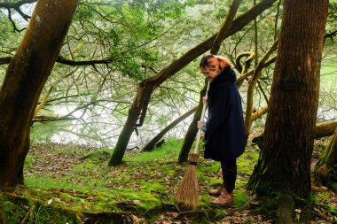 Cadı gibi giyinmiş bir kadın gölün yakınındaki bir ormanda yaprakları süpürüyor. Fantezi, gizem ve büyülü açık hava sahnesi kavramı