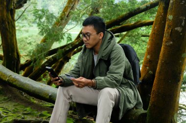 Orman tatili sırasında elinde kredi kartı olan bir adam ağaç gövdesinde otururken telefonuna bakıyor. Seyahat, dijital ödeme ve doğa kaçışı kavramı