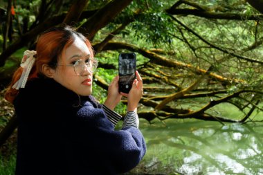 Seyahat ederken akıllı telefonuyla ormanın fotoğrafını çeken bir kadın. Doğa fotoğrafçılığı, seyahat yaşam tarzı ve açık hava keşfi kavramı.