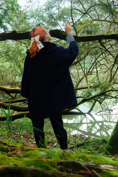 Göl kenarındaki bir ormanda yürürken ve yukarıya doğru bakan ceketli bir kadın. Macera kavramı, doğa keşfi ve açık hava merakı.