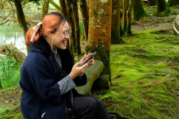 Ormanda bir ağaç gövdesinde oturan bir kadın tatildeyken telefonuyla mesaj atıyor. Seyahat kavramı, dijital yaşam tarzı ve doğa kaçışı..
