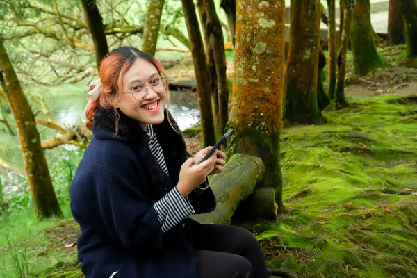 Elinde telefon tutan ve kameraya bakan, orman tatili sırasında ağaç gövdesinde oturan gülen bir kadın. Eğlence, seyahat ve dijital yaşam tarzı kavramı
