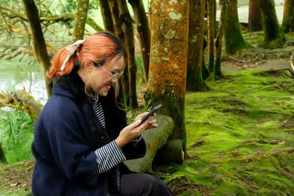 Ormanda bir ağaç gövdesinde oturan bir kadın tatildeyken telefonuyla mesaj atıyor. Seyahat kavramı, dijital yaşam tarzı ve doğa kaçışı..