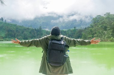 Sırt çantalı bir adam kollarını genişçe açar, sisli dağları ve yemyeşil ormanları olan yeşil bir göle bakar. Özgürlük, macera ve doğa keşfi kavramı