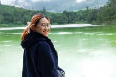 Dost canlısı Asyalı kadın tatildeyken göl kenarında kameraya gülümsüyor. Seyahat, mutluluk ve doğa rahatlığı kavramı