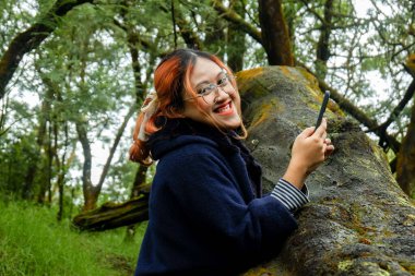 Ormanda tatildeyken elinde akıllı telefon tutan Asyalı kadın kameraya bakıyor. Seyahat kavramı, dijital yaşam tarzı ve doğa kaçışı.