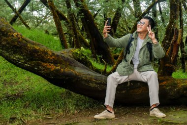 Asyalı bir adam ormanda tatildeyken cep telefonuyla selfie çekiyor.