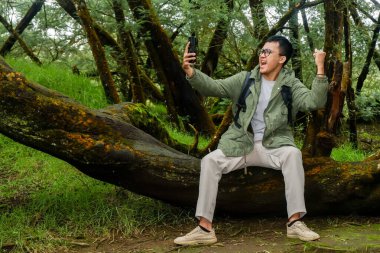 Asyalı bir adam ormanda tatildeyken cep telefonuyla selfie çekiyor.