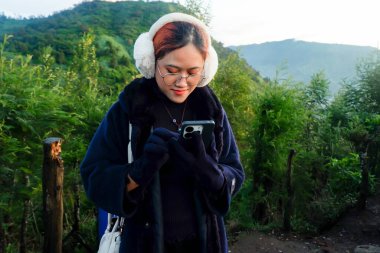 Bir tepede seyahat ederken akıllı telefon kullanan Asyalı bir kadın. Dijital yaşam tarzı, tatil, doğa ve açık hava rahatlama kavramı.
