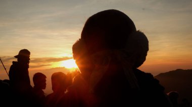 Sıcak güneş ışığı ve arka planda dramatik gökyüzü olan bir dağda gün doğumunu izleyen insanların siluetleri. Doğadaki barışçıl grup anı