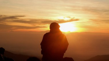 Puslu dağlar ve altın bir gökyüzü ile çevrili, parıldayan bir güneşin önünde duran bir insanın silueti. Barışçıl ve ilham verici açık hava anı