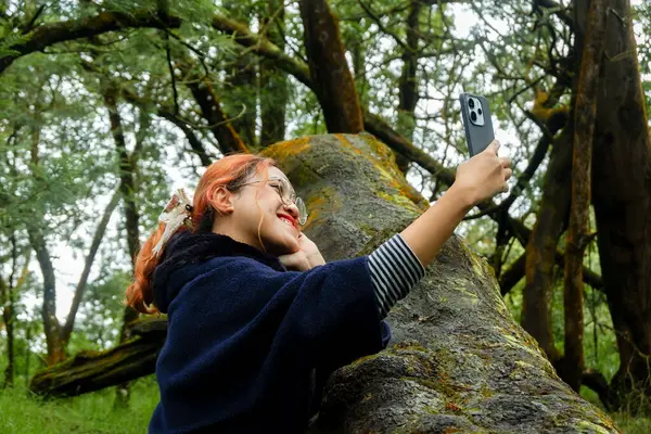 Asyalı bir kadın ormanda tatildeyken telefonuyla selfie çekiyor. Seyahat, doğa ve dijital yaşam tarzı kavramı