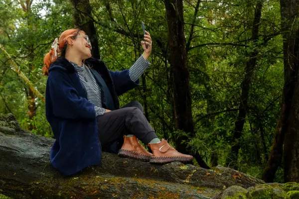 Asyalı bir kadın ormanda tatildeyken telefonuyla selfie çekiyor. Seyahat, doğa ve dijital yaşam tarzı kavramı